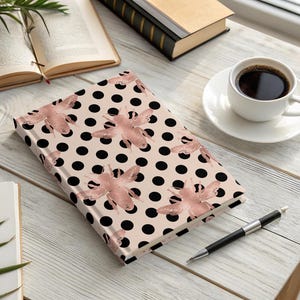 Op de afbeelding: Een hardcover notitieboek met een roze achtergrond, zwarte stippen en roze bijenillustraties. Het ligt op een houten tafel naast een kopje koffie, een pen en open boeken. Het boek is ongeveer 20 cm lang.