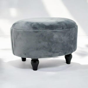 May include: A grey oval-shaped footstool with four black wooden legs. The footstool is upholstered in a soft, plush fabric.