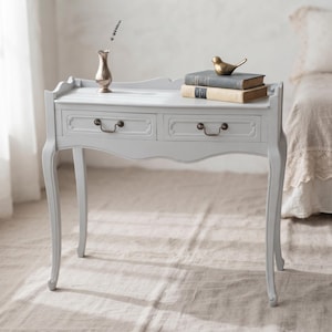May include: A light grey console table with two drawers, curved legs, and a scalloped top edge. The table features dark metal drawer pulls. On the surface sits a small silver vase with a sprig of lavender, two stacked books, and a brass bird figurine.