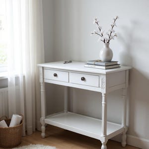 May include: A white wooden console table featuring two drawers with dark circular knobs, fluted legs, and a lower shelf. A white ceramic vase containing white floral branches sits atop two stacked books on the table surface.