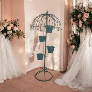 May include: A dark teal metal plant stand featuring a central pole topped with a decorative wire umbrella frame. Four matching teal cylindrical pots are attached at varying heights along the vertical pole, supported by a circular base.