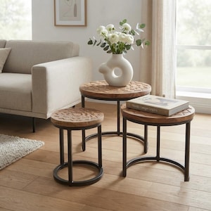 May include: A set of three round nesting tables with natural wood tops featuring carved geometric patterns and black metal frames. A white ceramic vase with white flowers sits on the tallest table, alongside a book titled Nordic Design Origins.