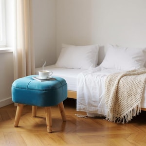 May include: A square, tufted teal velvet ottoman with four light-coloured wooden legs. A white ceramic mug filled with a steaming beverage sits on a small white saucer placed directly on the centre of the ottoman's flat, upholstered surface.