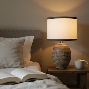 May include: A table lamp with a textured, dark brown, rounded ceramic base and a cylindrical white lampshade featuring thin black trim at the top and bottom edges. The lamp emits a warm, soft glow, illuminating the surrounding area.