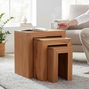May include: A set of three nesting tables crafted from light brown wood with a smooth finish. The tables are rectangular in shape and feature a clean, block-like design. They are stacked together in a descending size order, showcasing natural wood grain.
