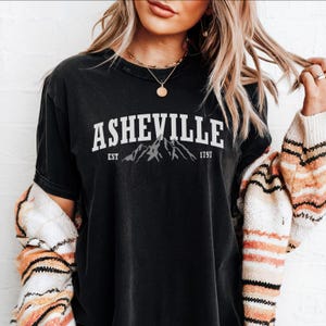 Pu&ograve; includere: T-shirt nera con la scritta "ASHEVILLE" in bianco sopra un disegno di montagne. Sotto, le parole "EST" e "1797". La maglietta &egrave; indossata con un cardigan fantasia.