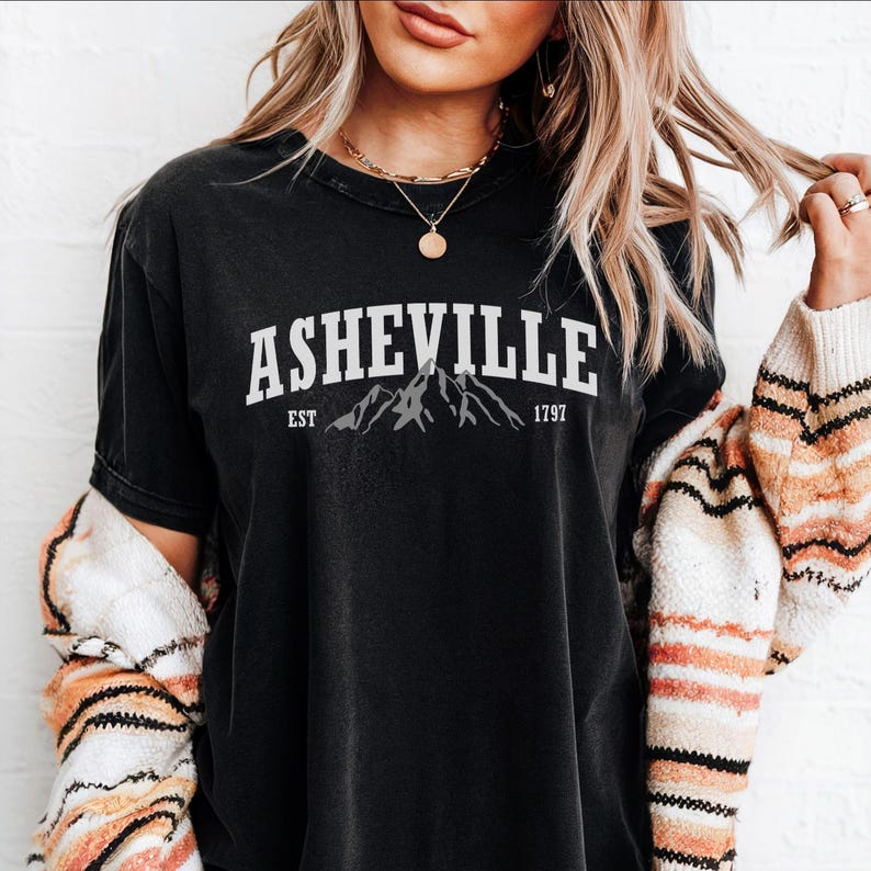 Pu&ograve; includere: T-shirt nera con la scritta "ASHEVILLE" in bianco sopra un disegno di montagne. Sotto, le parole "EST" e "1797". La maglietta &egrave; indossata con un cardigan fantasia.