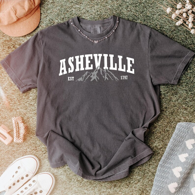 Pu&ograve; includere: T-shirt grigio scuro con la scritta "ASHEVILLE" in lettere bianche sopra una grafica di montagna. Sotto la grafica ci sono le parole "EST 1797". La maglietta &egrave; a maniche corte e ha uno scollo tondo. Una collana &egrave; intorno al collo.
