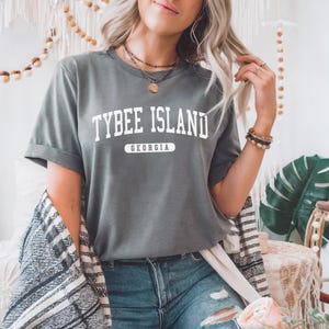 May include: A grey t-shirt with the words "TYBEE ISLAND GEORGIA" printed in white. The shirt has rolled-up sleeves and is worn with blue jeans. The style is casual and relaxed.