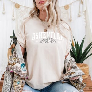 Pu&ograve; includere: Una maglietta color crema con la scritta "ASHEVILLE" in lettere bianche a blocchi sopra una grafica di catena montuosa. Il testo "EST 1797" &egrave; sotto. La maglietta fa parte di una collezione di abbigliamento casual.