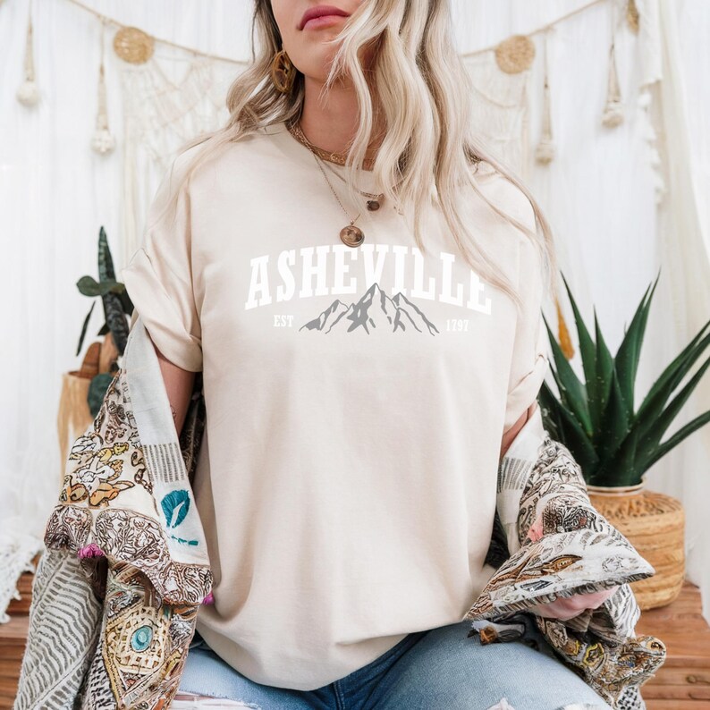 Pu&ograve; includere: Una maglietta color crema con la scritta "ASHEVILLE" in lettere bianche a blocchi sopra una grafica di catena montuosa. Il testo "EST 1797" &egrave; sotto. La maglietta fa parte di una collezione di abbigliamento casual.