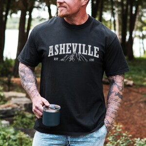 Pu&ograve; includere: T-shirt nera con la scritta "ASHEVILLE" in lettere bianche a blocchi sopra una grafica di montagna. Sotto la grafica ci sono le parole "EST" e "1797". La persona nell'immagine tiene in mano un bicchiere da viaggio blu scuro.