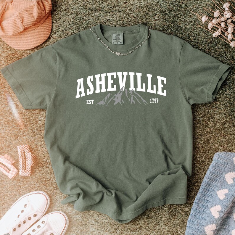 Pu&ograve; includere: T-shirt verde oliva con la scritta bianca "ASHEVILLE" sopra una grafica di montagna. Il testo "EST 1797" &egrave; sotto. La maglietta &egrave; presentata con accessori come un cappello rosa e scarpe da ginnastica bianche, suggerendo un look casual.