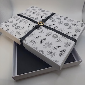 Peut inclure: Une boîte cadeau rectangulaire blanche avec un ruban noir et un emblème circulaire doré. Le couvercle présente un motif répétitif à l'encre noire composé d'insectes, de crânes, de papillons de nuit et de soleils. L'intérieur est noir.