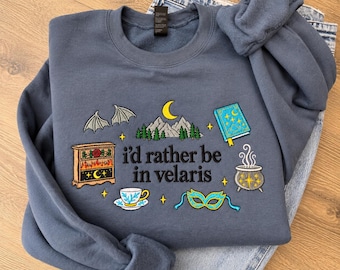 May include: A slate blue crewneck sweatshirt featuring embroidered designs including bat wings, a mountain range, a book, a fireplace, a teacup, a cauldron, and a mask. The text reads, "i'd rather be in velaris" in black lettering.