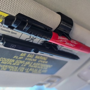 May include: A black plastic car visor clip with two writing utensils, a black pen and a red marker with the text "INKZALL" on it. The clip is attached to the visor of a car. The text on the visor reads "AIR BAG WARNING FLIP VISOR OVER COUSSIN GONFLABLE AVERTISSEMENT REPLIER LE PARESOLEIL ADVERTENCIA CON LA PARTE DE VIZA DE LA PARTE DE"
