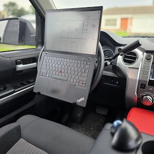 May include: A black ThinkPad laptop mounted on a steering wheel inside a vehicle. The laptop displays a technical diagram. The interior of the car is visible, including the dashboard, steering wheel, and a red-colored center console.