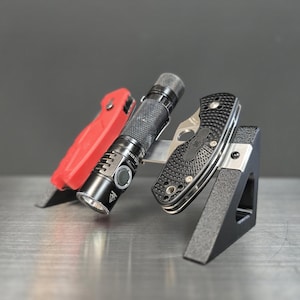 May include: A black metal EDC organizer holding a red folding knife, a black flashlight, and a black and silver pocket knife. The organizer is on a metallic surface with a gray background.