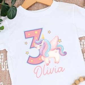 May include: White t-shirt with a pink number 3 and a unicorn with rainbow mane and tail. The unicorn is surrounded by stars. The name Olivia is printed below the unicorn.