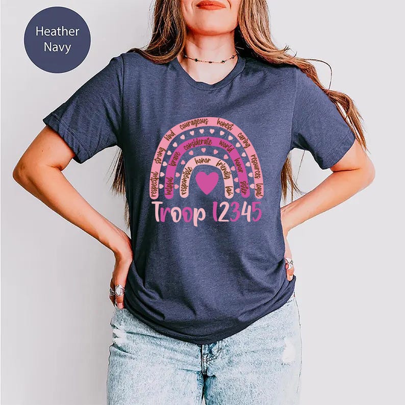 A woman wearing a navy blue t-shirt with a rainbow graphic and text, including the words "Heather Navy" and "Troop 12345".
