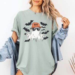 May include: A light green t-shirt featuring a cartoon ghost wearing sunglasses and an orange cap with the word "BOO". The ghost is surrounded by black bats, creating a Halloween theme.