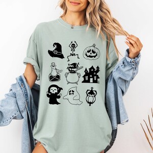 May include: A light green t-shirt featuring black Halloween-themed illustrations. The shirt displays a witch's hat, skeleton, jack-o'-lantern, potion bottle, cauldron, haunted house, grim reaper, ghost, and a decorative crown. The shirt is a comfortable fit.