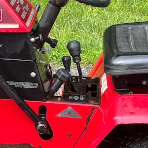May include: Close-up of a red and black riding lawn mower's control panel. The panel features multiple levers, a gear shift, and a black seat. The mower has a 6-speed transmission.