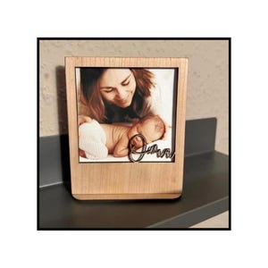 Könnte beinhalten: Ein Holzfotorahmen mit einem Foto von Mutter und Baby. Der Name "Jana" ist in Schreibschrift am unteren Rand des Fotos geschrieben. Der Rahmen ist rechteckig und steht auf einer dunklen Oberfläche.
