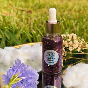 May include: A clear glass bottle with a gold dropper top, filled with a dark purple liquid and gold flakes. The bottle has a label that reads "blackfire shield protection oil." The image includes purple flowers and white cotton-like accents.
