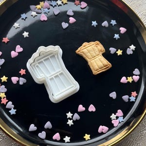 May include: A black plate with a gold rim, decorated with colourful sprinkles, featuring two cookie cutters shaped like coffee pots. One cutter is white plastic and the other is a baked cookie.