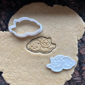 May include: Two white plastic turtle cookie cutters on a beige cookie dough surface. One cutter is on top of the dough, the other is next to it.