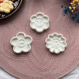 May include: Three white plastic flower-shaped cookie cutters with smiling faces are arranged on a pink knitted surface. The cutters have a daisy-like design. Baked cookies in the shape of flowers are on a black plate.