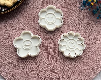 May include: Three white plastic flower-shaped cookie cutters with smiling faces are arranged on a pink knitted surface. The cutters have a daisy-like design. Baked cookies in the shape of flowers are on a black plate.