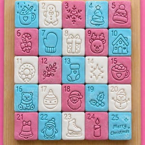 Può includere: Un set di venticinque piastrelle quadrate disposte in una griglia su una tavola di legno, con icone natalizie in rilievo e numeri da 1 a 25. Le piastrelle sono azzurre, bianche e rosa, con la scritta Merry Christmas sull'ultima piastrella.