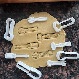 Puede incluir: Un conjunto de cortadores de galletas de plástico blanco con forma de diversas herramientas, incluyendo llaves inglesas, destornilladores y un martillo, están colocados sobre un trozo de masa extendida. La masa es de color marrón claro, y el fondo es una superficie oscura y moteada.