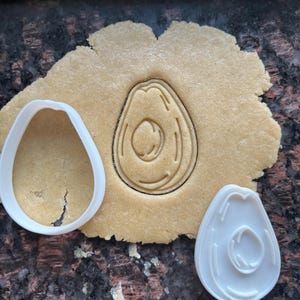 Puede incluir: Cortador de galletas de aguacate de plástico blanco sobre una superficie marrón con una masa de galletas que se ha cortado con el cortador.
