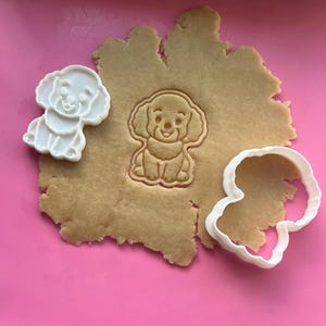 May include: Two white plastic cookie cutters shaped like a puppy with curly fur, one of which is pressed into a piece of light brown cookie dough, leaving a detailed embossed impression of the dog's face and body on the surface of the dough.