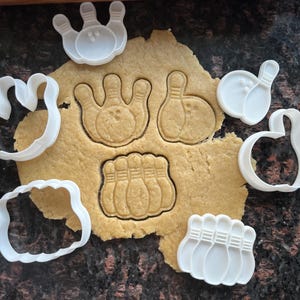 Puede incluir: Primer plano de masa para galletas con varios cortadores de galletas blancos con temática de bolos. La masa tiene impresiones de bolos y bolas de bolos. Un rodillo de madera está en el fondo. Los cortadores tienen forma de bolos y bolas de bolos.