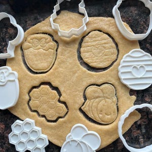 Könnte beinhalten: Nahaufnahme von Keksteig mit Ausstechformen zum Thema Bienen und weißen Kunststoff-Ausstechern. Die Ausstechformen umfassen eine Honigwabe, eine Biene und einen Bienenstock. Der Teig ist hellbraun und die Ausstecher sind weiß.
