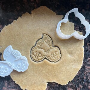 May include: A white plastic cookie cutter shaped like two cherries with skull faces, pressed into a flat piece of tan cookie dough, with a second identical white plastic cookie cutter resting on the dough surface nearby.