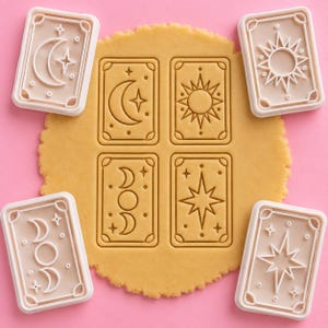 Pode incluir: Quatro cortadores de biscoitos retangulares com designs celestiais, incluindo uma lua crescente, um sol, uma fase lunar tripla e uma estrela. Os cortadores são bege claro e aparecem individualmente e pressionados em massa de biscoito.