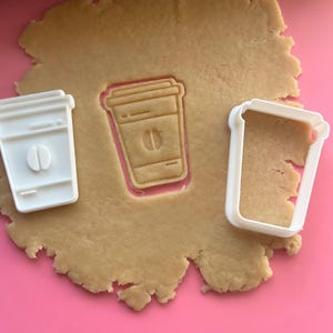 Könnte beinhalten: Ein Kaffeetassen-Ausstecher und ein ausgestochener Keks auf einer rosa Oberfläche. Der Ausstecher ist aus weißem Kunststoff und der Keks ist hellbraun. Der ausgestochene Keks hat ein Kaffeebohnen-Design.