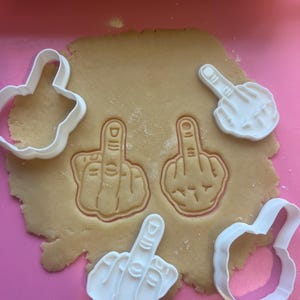 Op de afbeelding: Witte plastic koekjessnijders in de vorm van een hand die de middelvinger opsteekt, naast uitgesneden afdrukken in een deegvel. Het deeg ligt op een roze ondergrond.