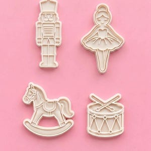 May include: A set of four off-white plastic cookie cutters featuring a nutcracker soldier, a ballerina in a tutu, a rocking horse, and a drum with two crossed drumsticks. Each cutter has a raised outline design for creating detailed impressions.