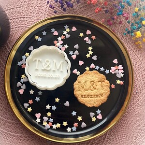 Peut inclure: Une assiette noire avec un bord doré contient un tampon à biscuits blanc avec les lettres T&M et une date, et un biscuit décoré avec les lettres M&T et la date 02.02.2026. L'assiette est parsemée de décorations en forme d'étoiles et de cœurs.
