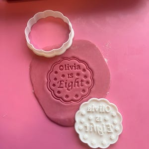 Może przedstawiać: Biała, karbowana wykrawaczka do ciastek i pasujący stempel z napisem "Olivia is Eight" znajdują się na różowej powierzchni z różowym ciastem. Drewniany wałek do ciasta znajduje się w tle. Stempel służy do odbijania tekstu w cieście.