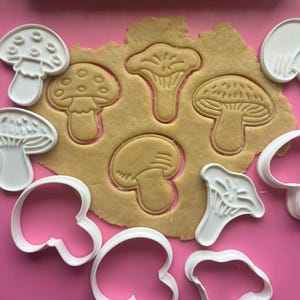 Op de afbeelding: Een set witte plastic koekjesvormers in de vorm van paddenstoelen, gerangschikt rond een stuk uitgerold beige koekjesdeeg. Het deeg bevat drie verschillende paddenstoelafdrukken, terwijl de vormers diverse schimmelvormen tonen.