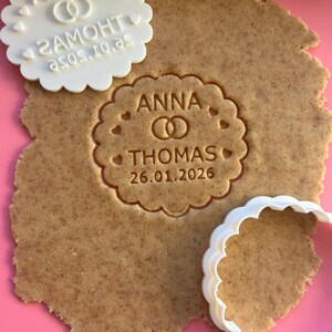 May include: A light brown, cut-out cookie with the names "ANNA" and "THOMAS" and the date "26.01.2026" imprinted on it. A white, scalloped-edge cookie cutter and a matching imprint tool are also present, along with a pink surface.