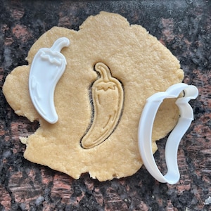 Puede incluir: Una imagen de primer plano de una masa con la impresión de un cortador de galletas de chile. Dos cortadores de galletas de chile de plástico blanco están junto a la masa. La masa es de color marrón claro y los cortadores de galletas son blancos.