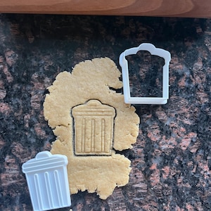 Może przedstawiać: Biała plastikowa foremka do ciastek w kształcie kosza na śmieci obok wyciętego ciastka w kształcie kosza na śmieci. Wałek do ciasta znajduje się w tle. Ciastko leży na ciemnej, nakrapianej powierzchni.